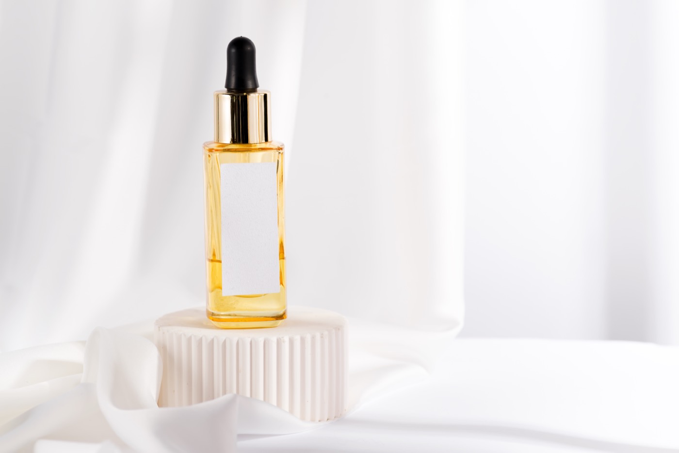 Sérum d'Or — a golden facial oil in a glass dropper bottle on a fluted stone podium with silk.